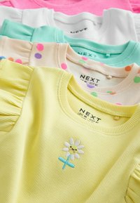 Pila de cinco camisetas para bebé con textura acanalada en amarillo, lunares, verde menta, blanco y rosa; la camiseta amarilla tiene una margarita sonriente bordada.