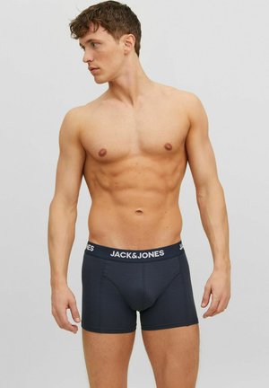 Homme torse nu aux cheveux courts et bouclés portant des boxers bleu marine Jack & Jones, debout devant un fond gris uni.