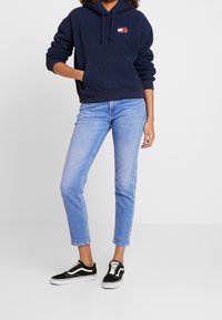 Felpa in pile blu navy con tasca frontale e logo, abbinata a jeans dritti azzurri e sneakers nere con dettagli bianchi.