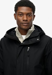 Zwarte waterdichte jas met een capuchon, voorzien van een ritszak op de borst, een logo aan de linkerkant, en laag over een lichtgrijze trui.