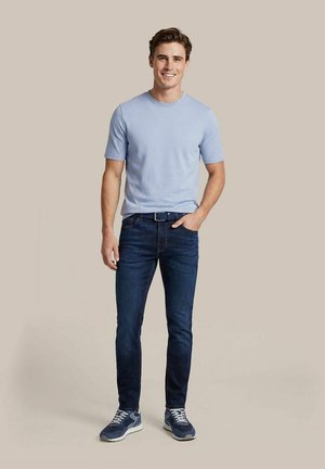 Jonge man staat glimlachend, draagt een lichtblauw shirt met korte mouwen, donkerblauwe jeans, blauwe riem en blauwe sneakers, beige achtergrond.