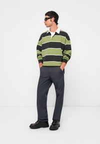 Uomo che indossa occhiali da sole neri, una maglietta a righe verdi e nere con maniche lunghe da rugby, pantaloni grigio scuro e scarpe nere spesse, in piedi con le mani in tasca.