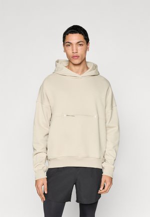 Beige sweatshirt met capuchon, voorzien van een voorzak met rits en geribde boorden. Gestyled met donkere korte broek, tegen een effen achtergrond.