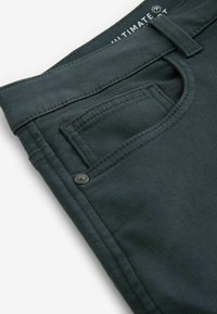 Pantaloni verzi închis dintr-un material ușor texturat, având un buzunar frontal și un detaliu din metal în partea de sus a buzunarului.
