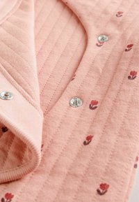 Lyst lyserød ribbet stof med små blomsterprint. Har metalliske trykknapper og en foldet kravedesign. Blød tekstur.