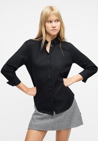 Νεαρή γυναίκα με ξανθά μαλλιά που φοράει μαύρο πουκάμισο με κουμπιά και ασπρόμαυρη καρό φούστα, με τα χέρια στη μέση, κοιτάζει προς το πλάι.