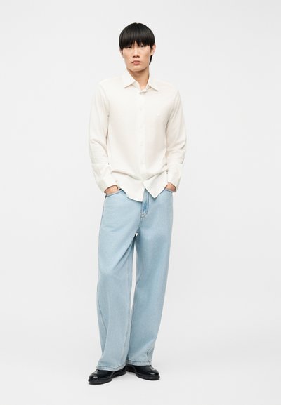 Νεαρός άνδρας με μαύρα μαλλιά που φοράει λευκό πουκάμισο με μακριά μανίκια μέσα από ανοιχτόχρωμα φαρδιά τζιν παντελόνια, με τα χέρια στις τσέπες και μαύρα παπούτσια.