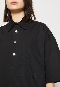 Czarna koszula z krótkim rękawem wykonana z lekkiego materiału. Wyposażona w kołnierz zapinany na guziki, kieszeń na piersi oraz guziki w srebrnym kolorze.