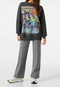 Sudadera negra con estampado gráfico colorido que presenta motocicletas y texto; combinada con pantalones de pierna ancha grises y zapatillas ligeras.