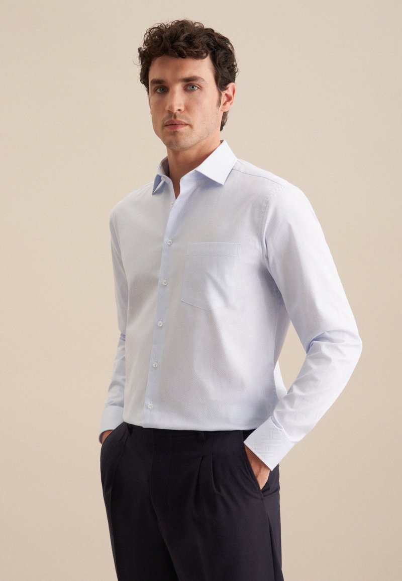 Chemise bleu clair à manches longues avec une petite poche, ornée d'un motif géométrique subtil, assortie à un pantalon sombre.