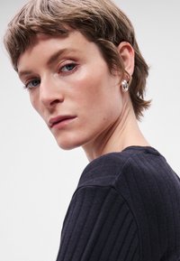 Κοντινό πλάνο ενό�ς ατόμου με κοντά μαλλιά που φοράει ένα σκούρο ριπτό μπλουζάκι. Είναι ορατά εντυπωσιακά ασημένια σκουλαρίκια. Το φόντο είναι απλό και φωτεινό.