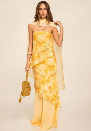 Mujer con vestido amarillo sin tirantes con estampado floral y capa transparente, sosteniendo un bolso amarillo tejido sobre un fondo beige liso.