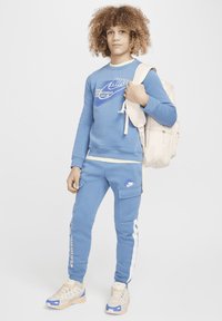 Sudadera Nike de color azul claro con logo, pantalones jogger a juego con franjas laterales blancas y zapatillas beige con acentos coloridos. Mochila en mano.