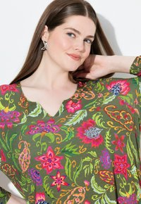 Groene bloemenblouse met een V-hals ontwerp, versierd met levendige roze, rode en blauwe bloemen, en ingewikkelde bladerenpatronen. Zachte stofstructuur.