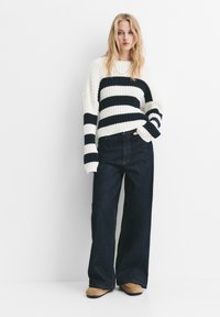 Blonde vrouw in een wit en marineblauw gestreept sweater, met wijde donkere jeans en tan kleurige moccasinschoenen, staande tegen een witte muur.
