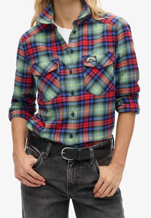Persona que lleva una camisa de cuadros roja, azul y verde con botones y mangas remangadas, metida dentro de unos vaqueros negros con un cinturón negro.