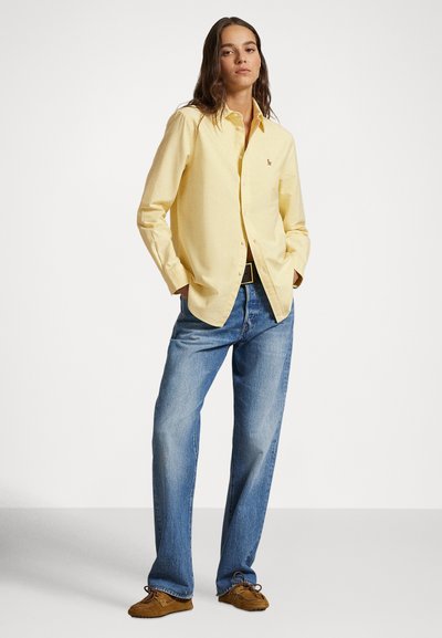 Dzeltena ar pogām aizpogājama krekla, ar garām piedurknēm un brīvu piegriezumu, kombinēta ar gaismzilām vaļīgām džinsu biksēm un brūnām mokasīnu kurpēm.