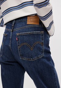 Σκούρα μπλε τζιν από τζιν με κλασικό σχέδιο, διακοσμημένα με δερμάτινο ταμπελάκι και κίτρινη ραφή. Δύο πίσω τσέπες με εμβληματική λεπτομέρεια ραφής.