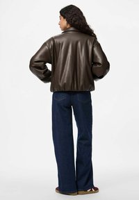 Femme aux longs cheveux bouclés portant une veste en cuir brun foncé, un jean bleu large et des baskets rouges, se tenant les mains sur les hanches et faisant face à l'extérieur.