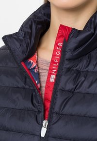 Marinblå puffjacka med hög krage; har ett röd innerfoder med blommönster och en vit dragkedja med logodetalj på framsidan.