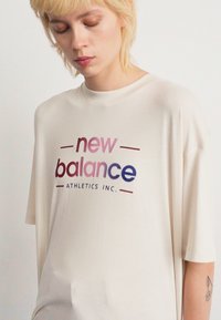 Personne aux cheveux blonds courts, regardant vers le bas, portant un T-shirt oversize de couleur écrue avec le texte "new balance ATHLETICS INC." en dégradé de rose à bleu.