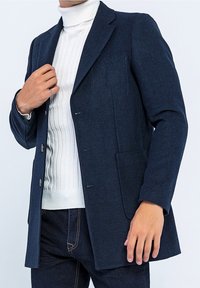 Navy ullblandad kappa med två framfickor, en enkelknäppt design och slät textur, parat med en vit ribbad polotröja.