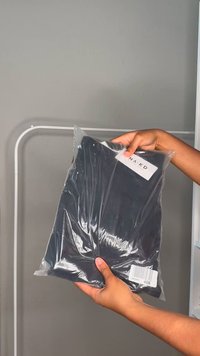 Juodas drabužis supakuotas skaidrioje plastikinėje maišelėje. Ant šono yra etiketė su užrašu "NA-KD" ir brūkšninis kodas.