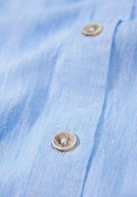 Jasnoniebieska tkanina o lekko teksturowanej powierzchni. Posiada okrągły, beżowy guzik z subtelnym wzorem, umieszczony po lewej stronie.