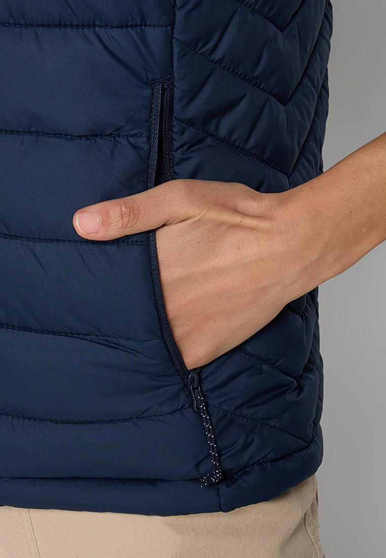 Main dans une poche zippée sur le côté d'un gilet matelassé bleu marine porté sur un pantalon beige.