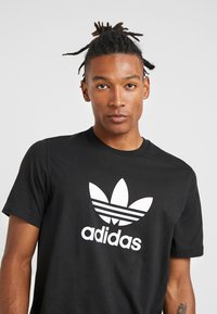 Fekete pamutt póló, nagyméretű fehér Adidas trefoil logóval és felirattal. Rövid ujjú, kerek nyakkivágású, sima textúrájú.