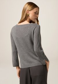 Grauer Strickpullover mit Boot-Ausschnitt, Dreiviertelärmeln und seitlichen Schlitzen am Saum. Getragen mit dunklen, maßgeschneiderten Hosen.