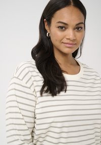 White long-sleeve shirt with gray horizontal stripes, round neckline, and soft texture. Model has dark hair and wears silver hoop earrings.