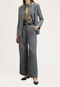 Costume gris sur mesure avec pantalon à jambes larges et blazer élégant. En dessous, un chemisier imprimé léopard avec un col fendu. Ceinture noire et chaussures noires.