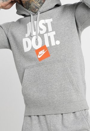 Grauer Nike-Hoodie mit weißem "JUST DO IT."-Text und einem orangefarbenen Nike-Label, getragen von einer tätowierten Person, die die Kapuze anpasst.