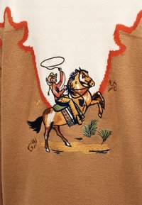 Καουμπόι με λασσό που καβαλάει ένα άλογο που σηκώνεται στα δύο, κεντημένο σε καφέ ύφασμα με λευκές και κόκκινες φλόγες ως διακοσμητικά περιθώρια.