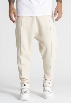 Mężczyzna ubrany w kremowe, teksturowane spodnie dresowe i białe trampki za kostkę z beżowymi sznurowadłami, stojący na jednolitym tle.