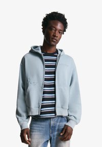 Sweatshirt à capuche bleu clair avec une fermeture éclair à l'avant et deux poches, porté sur une chemise rayée noir et blanc. Tissu doux avec une coupe décontractée.