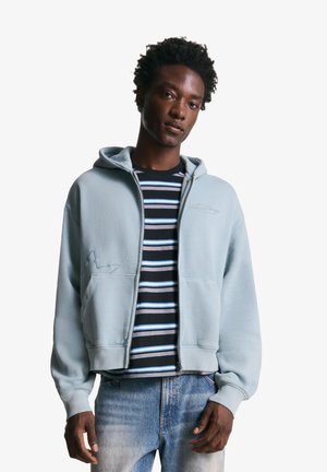 Sweatshirt à capuche bleu clair avec une fermeture éclair à l'avant et deux poches, porté sur une chemise rayée noir et blanc. Tissu doux avec une coupe décontractée.