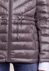 Kviltad dunjacka i en dämpad grå färg, med hög krage, dragsko i midjan, sidofickor och metallisk dragkedjestängning.