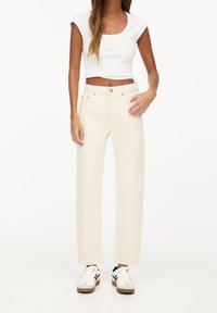 Jean beige à jambes larges avec une taille haute, cinq poches et une fermeture à bouton, associé à un crop top blanc ajusté et des baskets.