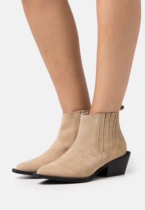 Beige ruskind støvletter med spids tå, teksturerede sidepaneler og lav blokhæl. Designet har en glat finish og minimale detaljer.