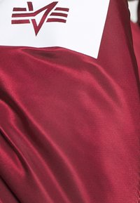 Tessuto in satin color maroon con una texture liscia, caratterizzato da un pannello bianco e un design geometrico di logo maroon sopra.