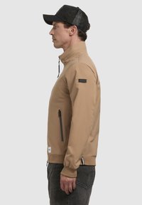 Chaqueta marrón con un diseño ajustado, cuello alto y puños elásticos. Presenta un bolsillo con cremallera y una etiqueta de la marca en la manga. Combinada con una gorra negra.