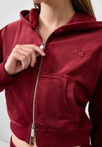 Burgunderfarbene Zip-Hoodie aus weichem Material mit einer Fronttasche, gerippten Bündchen und einem gestickten Logo mit Flügeln auf der oberen linken Seite.