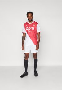 Camiseta corta de mangas rojas y blancas con un diseño diagonal, que presenta un logo y cuello, combinada con pantalones cortos blancos y calcetas y botas negras.