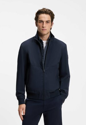 Jeune homme portant une veste zippée bleu marine par-dessus une chemise à carreaux et un sous-vêtement blanc, debout avec une main dans la poche.
