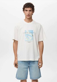 Hvid bomulds t-shirt med et blåt og hvidt grafisk motiv af måger med teksten "fanget af havet." Kombineret med lyseblå denim shorts.