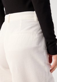 Pantalon en velours côtelé blanc avec texture à côtes verticales, doté d'une poche arrière et d'une taille haute. Haut noir à manches longues partiellement visible.