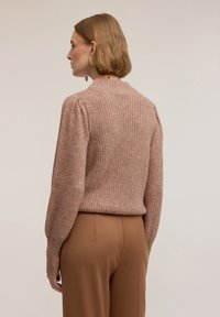 Różowy sweter na drutach z teksturowanym wzorem, okrągłym dekoltem i prążkowanym dołem, zestawiony z brązowymi spodniami. Miękki, luźny krój.