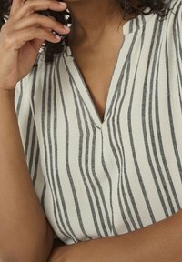 Gestreepte blouse met een v-hals, voorzien van verticale zwart-witte strepen, gemaakt van een gestructureerde stof. Close-up van de kraag en hand.